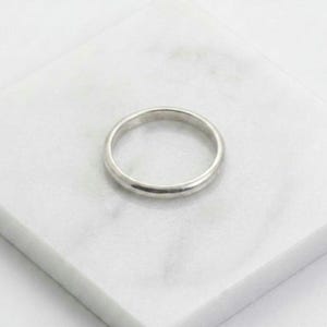 May include: A silver band ring with a smooth, rounded design. The ring is displayed on a white, marble-like surface. The ring's simple, classic style makes it suitable for everyday wear.
