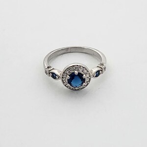Puede incluir: Un anillo de plata con una piedra preciosa azul rodeada de piedras blancas m&aacute;s peque&ntilde;as. El anillo tiene dos piedras azules m&aacute;s peque&ntilde;as a cada lado de la piedra central.