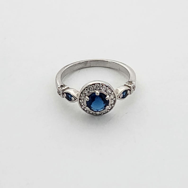 Puede incluir: Un anillo de plata con una piedra preciosa azul rodeada de piedras blancas m&aacute;s peque&ntilde;as. El anillo tiene dos piedras azules m&aacute;s peque&ntilde;as a cada lado de la piedra central.