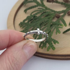 Pode incluir: Um anel de prata com um design de cruz. O anel é segurado em uma mão, mostrando seus detalhes intrincados. A cruz está centralizada no anel, com elementos decorativos ao longo da banda. O anel está em uma superfície de madeira com folhagem verde ao fundo.