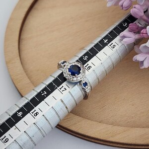 Puede incluir: Un anillo de plata con una gran piedra preciosa azul rodeada de piedras blancas m&aacute;s peque&ntilde;as. El anillo est&aacute; dimensionado en un medidor de anillos que muestra una talla 16.