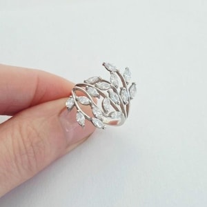 May include: A silver ring featuring a leaf design. The ring has multiple curved bands with small, marquise-cut, clear gemstones set along the bands, resembling leaves. The ring is held against a white background.