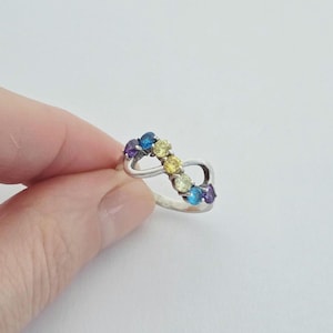 May include: A silver-colored ring in an infinity symbol design, adorned with a series of colorful gemstones. The gemstones are in shades of purple, blue, and yellow. The ring is held by a person's hand against a white background.