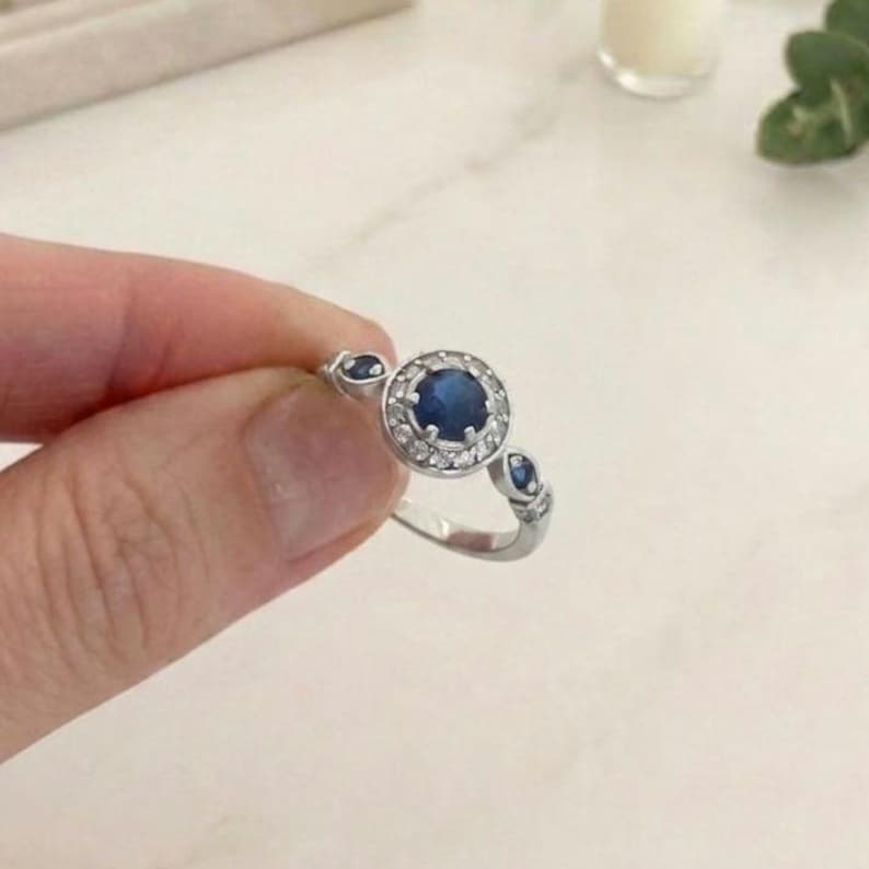 Close-up photograph focusing on a vintage-style halo ring. The sterling silver ring features a deep blue round lab-created sapphire at its center, encircled by a halo of small white cubic zirconia stones. Small blue sapphire accents are visible on the split-shank band. The ring is held gently by a hand against a clean, bright white marble surface.