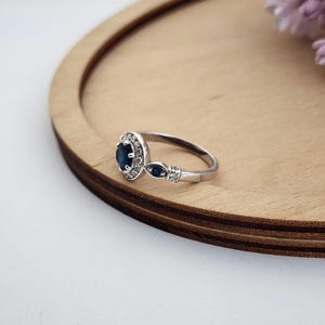 Puede incluir: Un anillo de plata con un zafiro azul central rodeado de peque&ntilde;os diamantes blancos. El anillo tiene un dise&ntilde;o delicado con dos zafiros azules m&aacute;s peque&ntilde;os en la banda.