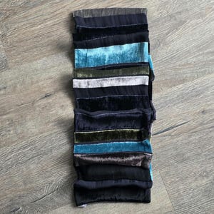 May include: A vertical stack of fabric strips in teal, black, silver, and olive green. The strips, likely velvet, create a textured look. The arrangement suggests a modern design. The fabric strips are of varying widths, adding visual interest to the piece.