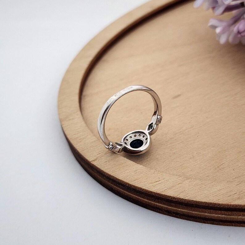 Puede incluir: Un anillo de plata con una piedra ovalada azul oscuro rodeada de piedras m&aacute;s peque&ntilde;as transparentes. El anillo est&aacute; sobre una superficie de madera.