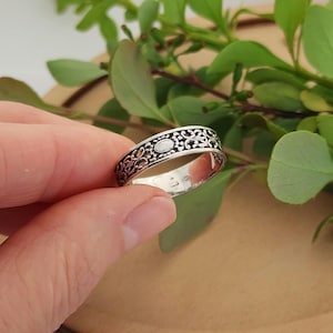Puede incluir: Un anillo de plata con un intrincado diseño floral. El anillo presenta un centro ovalado y patrones calados detallados. El anillo se sostiene en una mano, con hojas verdes en el fondo.