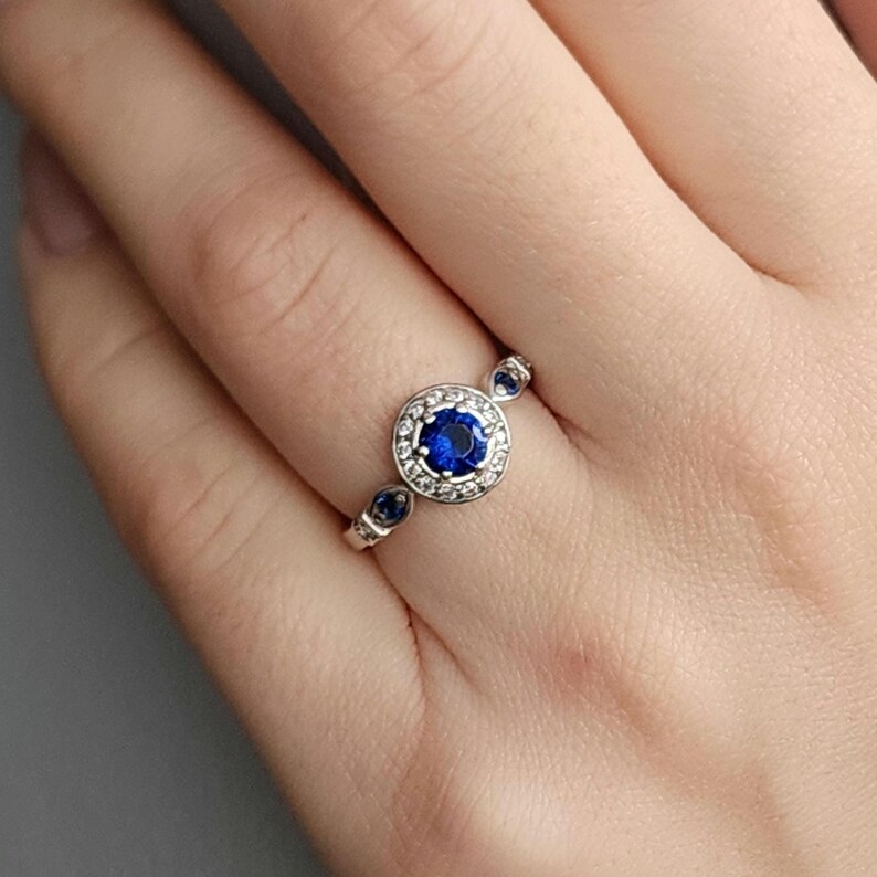 Puede incluir: Un anillo de plata con un zafiro azul central rodeado de peque&ntilde;os diamantes blancos. El anillo tiene tres peque&ntilde;os zafiros azules en la banda.
