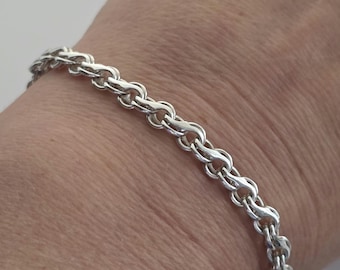 Puede incluir: Un brazalete plateado con un diseño detallado de eslabones de cadena. El brazalete está hecho de un metal brillante y se muestra en una muñeca. Los eslabones están estrechamente conectados, creando una apariencia texturizada. El brazalete mide unos 19 cm de largo.