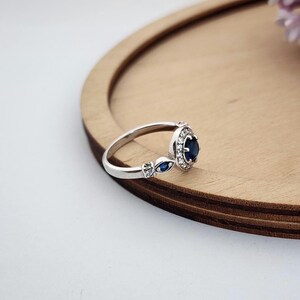 Puede incluir: Un anillo de plata con una gran piedra preciosa azul rodeada de piedras preciosas blancas m&aacute;s peque&ntilde;as. El anillo tiene dos piedras preciosas azules m&aacute;s peque&ntilde;as a cada lado de la piedra principal.