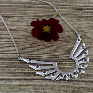 May include: A silver necklace with a geometric design featuring amethyst gemstones. The necklace is in the shape of wings with a rounded bottom.