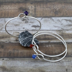 May include: Two silver bangle bracelets with gemstone accents. The top bracelet has a purple gemstone, the bottom bracelet has a red gemstone and a blue gemstone.