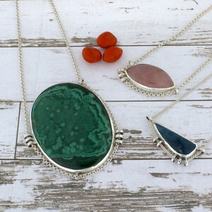 Puede incluir: Tres collares de plata con diferentes colgantes de piedras preciosas. El primer collar tiene un gran colgante ovalado de malaquita verde. El segundo collar tiene un colgante ovalado rosa de cuarzo rosa. El tercer collar tiene un colgante triangular azul.