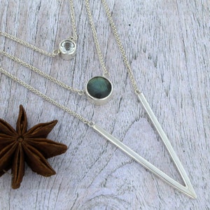 May include: A silver necklace with three strands and two pendants. The larger pendant is a round, dark green labradorite stone. The smaller pendant is a clear, round stone. The necklace has a V-shaped pendant.