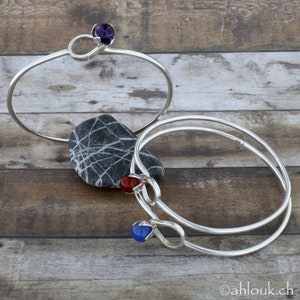 May include: Two silver bangle bracelets with gemstone accents. The top bracelet features a purple gemstone, the bottom bracelet features a red gemstone and a blue gemstone.