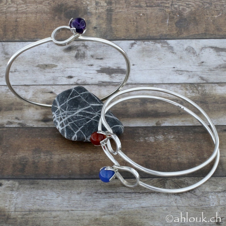 May include: Two silver bangle bracelets with gemstone accents. The top bracelet features a purple gemstone, the bottom bracelet features a red gemstone and a blue gemstone.