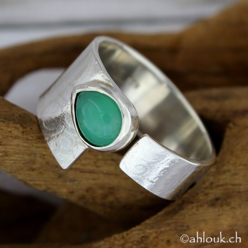 May include: A silver ring with a green gemstone set in a textured band. The ring is open on one side and has a textured finish.