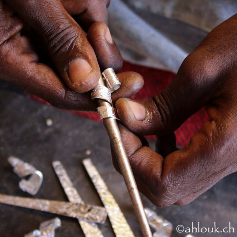 May include: A person's hands are shown holding a silver metal rod with multiple rings attached to it. The rings are decorated with intricate patterns. The person is working on the rings, possibly making jewelry.