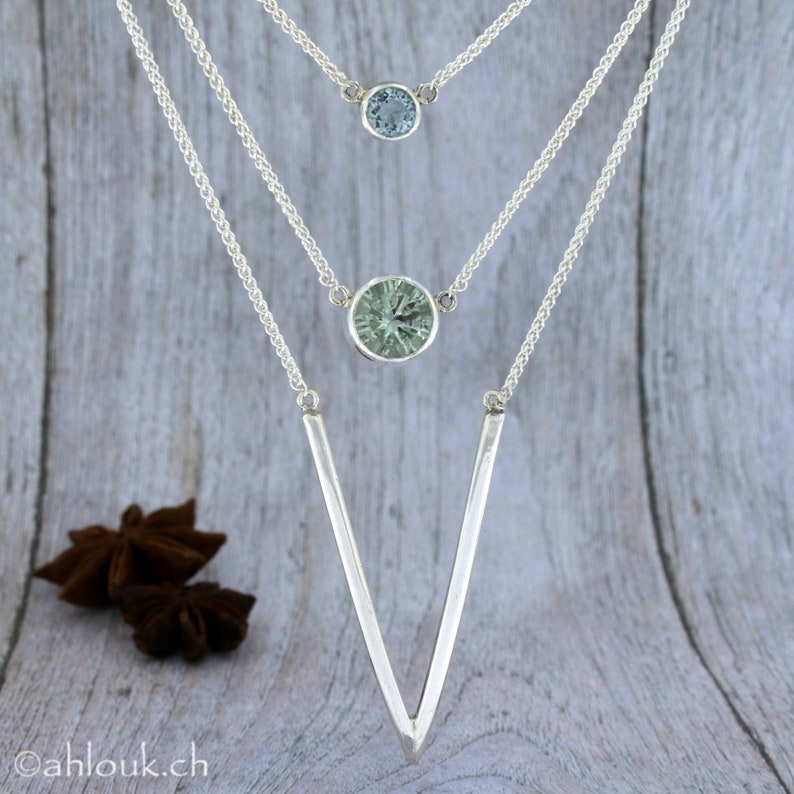 May include: A silver necklace with three chains and two gemstone pendants. The top pendant is a small, light blue gemstone. The bottom pendant is a larger, green gemstone. The chains are connected to a V-shaped silver pendant.