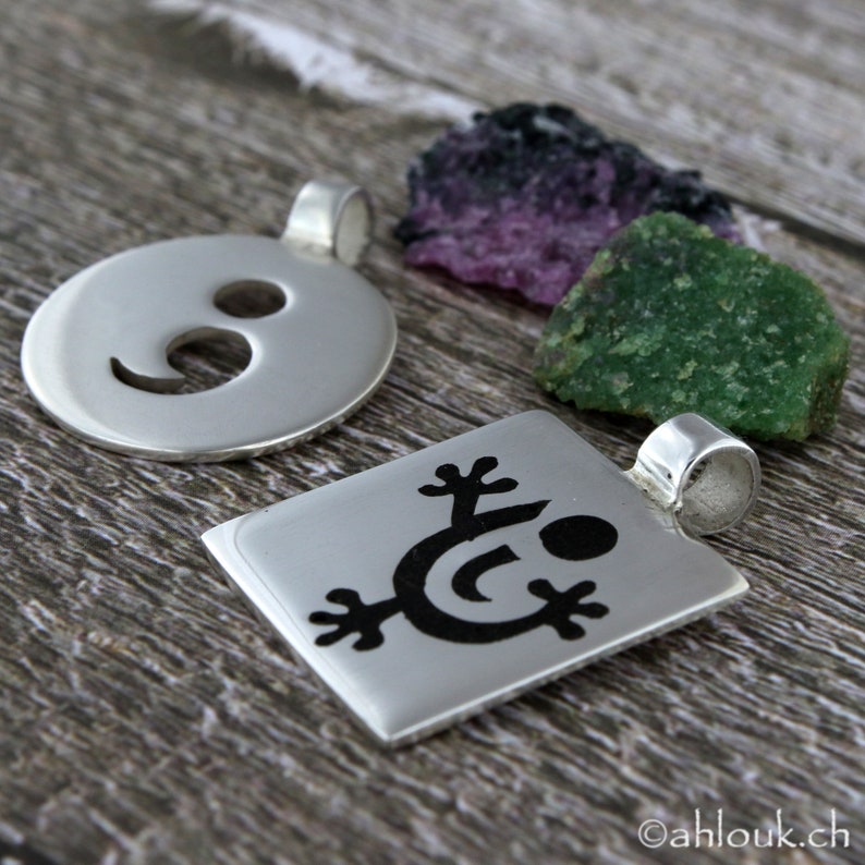 May include: Two silver pendants with different designs. One pendant is round with a semi-circle cut out. The other pendant is square with a stylized figure of a person with arms raised.