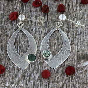 May include: A pair of silver earrings with a green stone set in each. The earrings are shaped like a teardrop with a cutout in the center. The earrings are on a wooden surface.