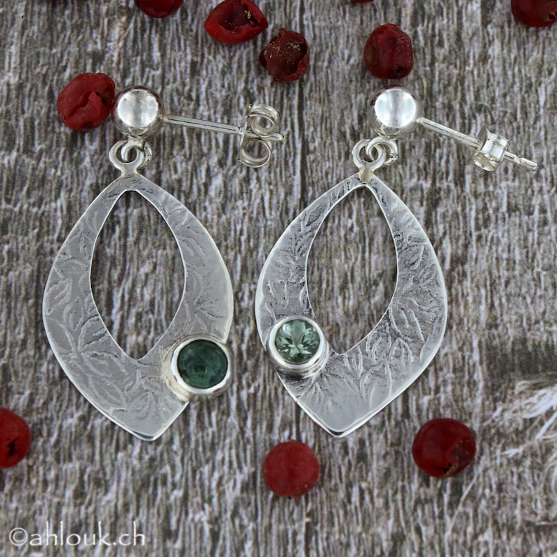 May include: A pair of silver earrings with a green stone set in each. The earrings are shaped like a teardrop with a cutout in the center. The earrings are on a wooden surface.