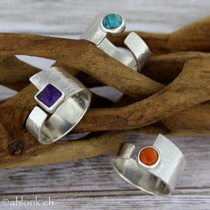 May include: Three silver rings with different colored gemstones. The rings are on a wooden surface. The gemstones are turquoise, amethyst, and carnelian.