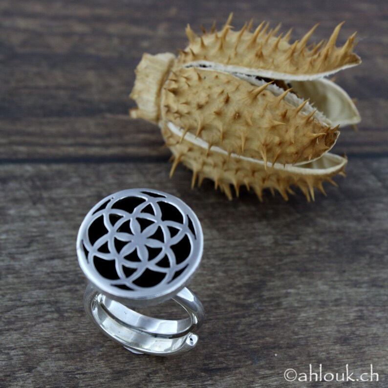 May include: A silver ring with a circular design featuring a flower of life pattern. The ring is on a wooden surface.