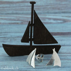 K&ouml;nnte beinhalten: Ein Paar silberne Segelschiff-Ohrringe mit strukturierter Oberfl&auml;che. Die Ohrringe werden auf einem Holzmodell eines Segelschiffs pr&auml;sentiert.