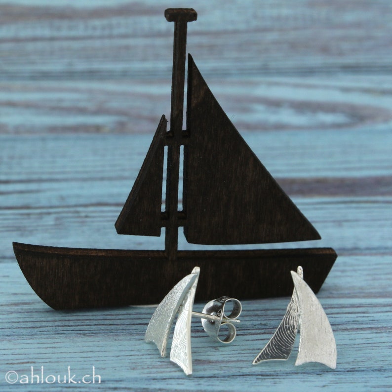 K&ouml;nnte beinhalten: Ein Paar silberne Segelschiff-Ohrringe mit strukturierter Oberfl&auml;che. Die Ohrringe werden auf einem Holzmodell eines Segelschiffs pr&auml;sentiert.