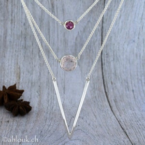 May include: A silver necklace with three chains and two gemstone pendants. The top pendant is a small, round, dark pink gemstone. The bottom pendant is a larger, round, light pink gemstone. The chains are connected to a V-shaped silver pendant.