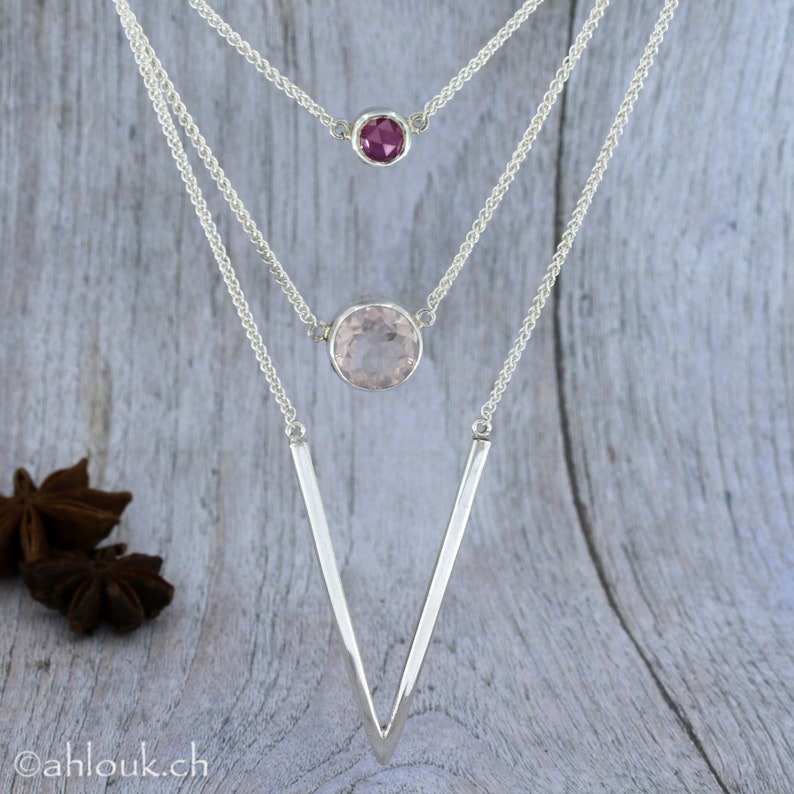 May include: A silver necklace with three chains and two gemstone pendants. The top pendant is a small, round, dark pink gemstone. The bottom pendant is a larger, round, light pink gemstone. The chains are connected to a V-shaped silver pendant.