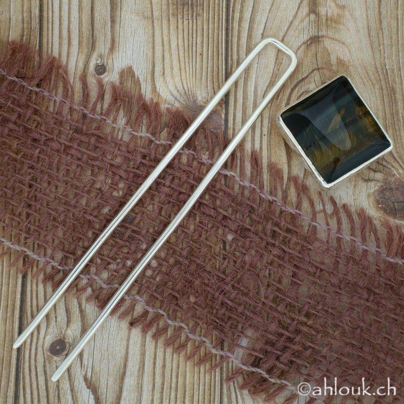 May include: Silver hair stick with a square, brown and gold gemstone. The stick is lying on a brown burlap fabric.