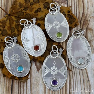 May include: A collection of six silver pendants with oval shapes and intricate geometric designs. Each pendant features a different colored gemstone: turquoise, red, green, amethyst, and a light blue stone. The pendants are displayed on a wooden surface with a leaf.