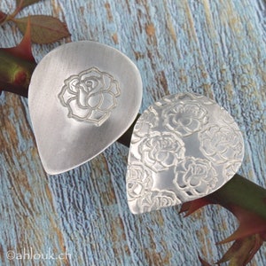 May include: Two silver teardrop-shaped pendants with a rose design. One pendant features a single rose in the center, while the other pendant is covered in a repeating rose pattern.