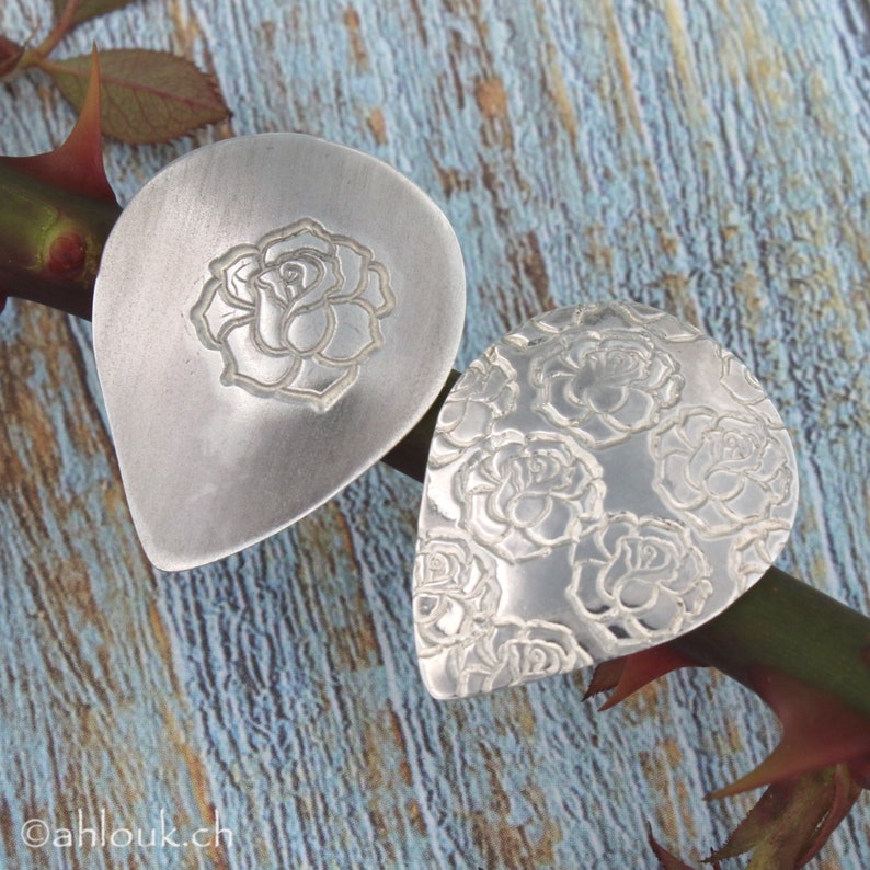 May include: Two silver teardrop-shaped pendants with a rose design. One pendant features a single rose in the center, while the other pendant is covered in a repeating rose pattern.