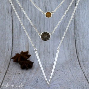 May include: A silver necklace with three pendants. The top pendant is a small, round, light brown gemstone. The middle pendant is a larger, round, dark brown gemstone. The bottom pendant is a V-shaped piece of silver.