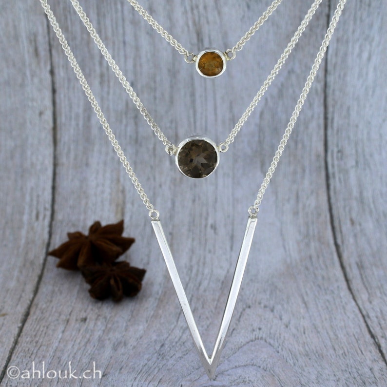May include: A silver necklace with three pendants. The top pendant is a small, round, light brown gemstone. The middle pendant is a larger, round, dark brown gemstone. The bottom pendant is a V-shaped piece of silver.