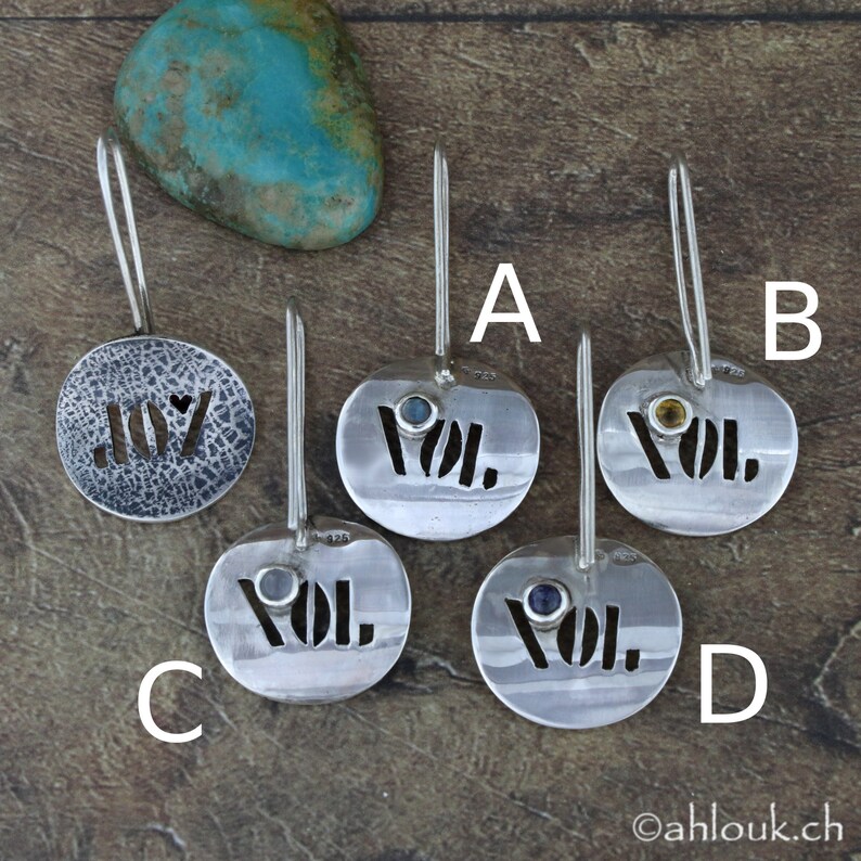 Puede incluir: Cinco colgantes de plata con diferentes dise&ntilde;os. Un colgante dice "JOY" con un recorte en forma de coraz&oacute;n. Los otros cuatro colgantes dicen "VOL." con una piedra preciosa de color diferente en cada uno. Los colgantes est&aacute;n sobre una superficie de madera.