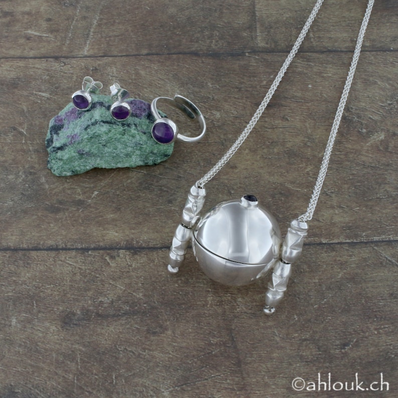 May include: A silver necklace with a round pendant that opens. The pendant has a small purple gemstone on top. The necklace is on a wooden surface with a pair of earrings and a ring with purple gemstones.