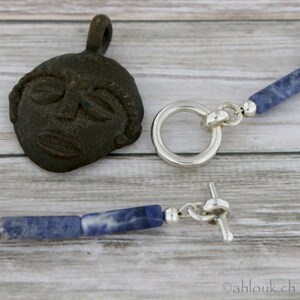 May include: A silver necklace with a blue stone pendant and a silver toggle clasp. The pendant is a rectangular shape with a blue stone that has white veins running through it. The necklace is on a wooden surface.