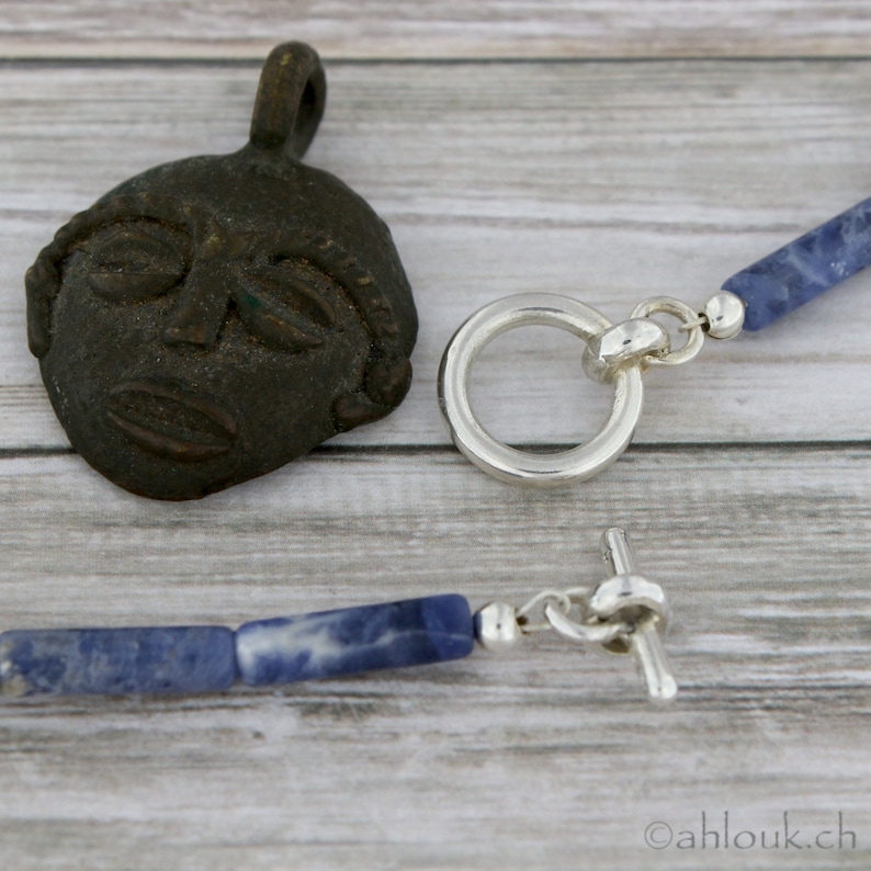 May include: A silver necklace with a blue stone pendant and a silver toggle clasp. The pendant is a rectangular shape with a blue stone that has white veins running through it. The necklace is on a wooden surface.