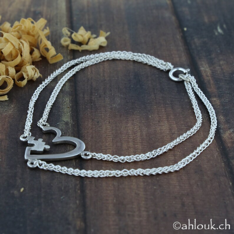 May include: A silver necklace with a heart-shaped pendant featuring two interlocking puzzle pieces. The necklace is made of two strands of chain.