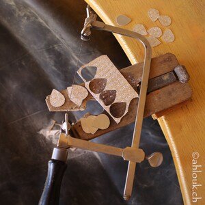 May include: A silver-toned jewelry saw with a wooden base is used to cut out a sheet of metal. The metal sheet has multiple teardrop shapes cut out of it.