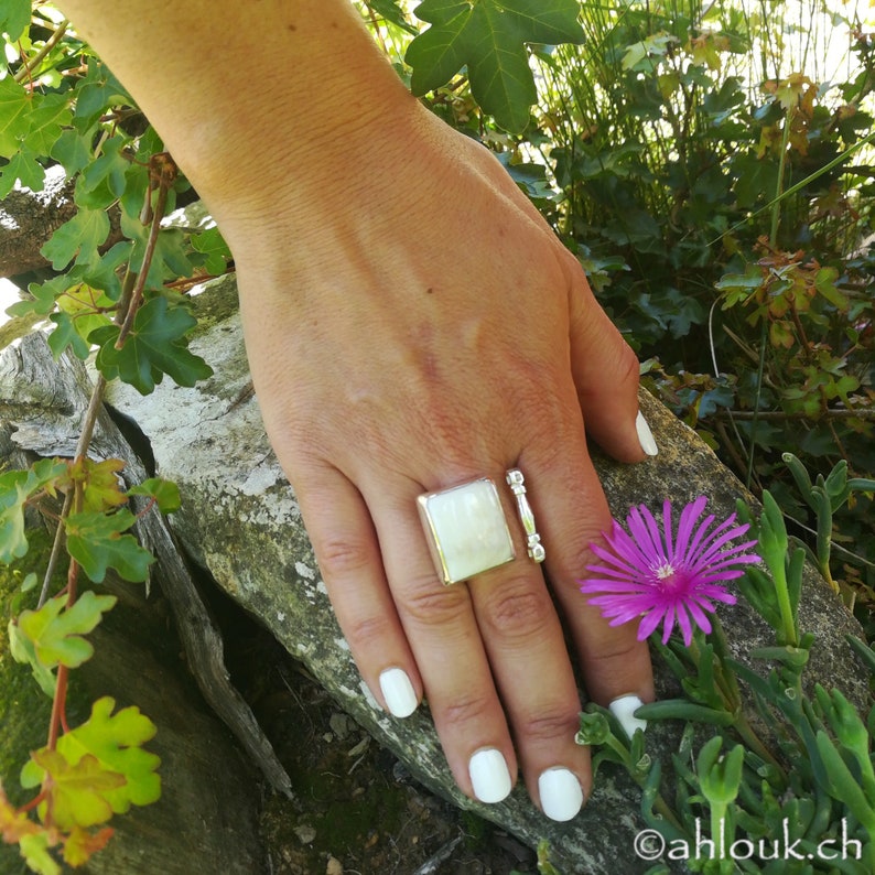 May include: A silver ring with a white square stone set in the centre. The ring is worn on the index finger of a hand with white nail polish.
