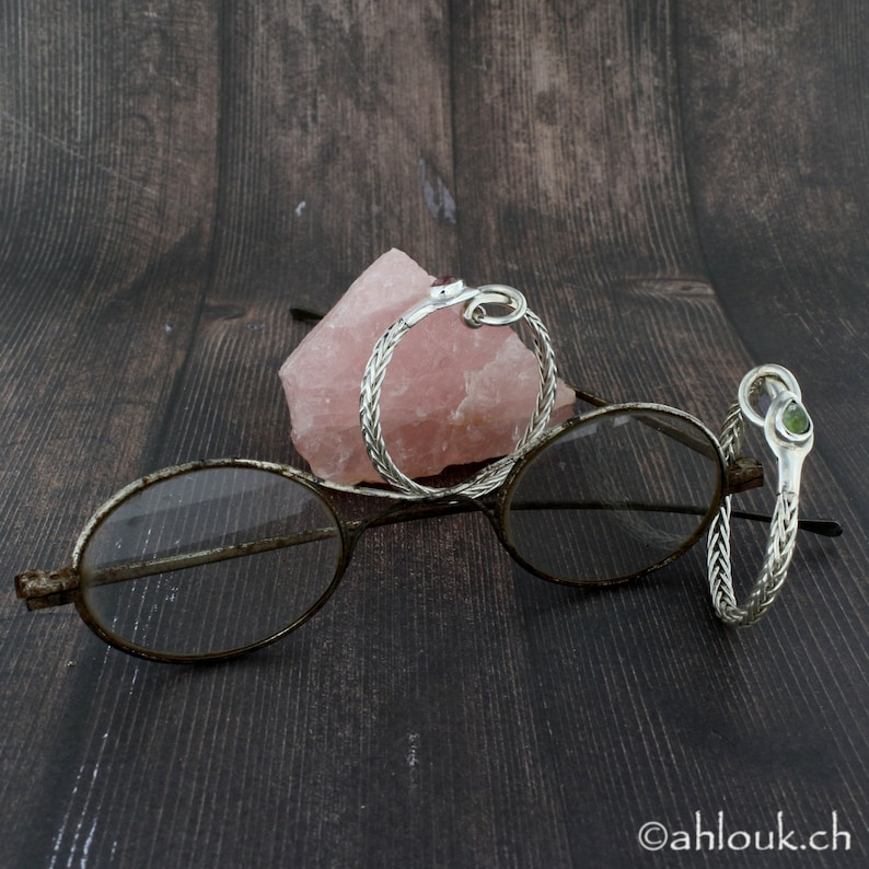 May include: Two silver bracelets with braided designs, one with a green gemstone and the other with a pink gemstone, resting on a pink crystal and a pair of eyeglasses.