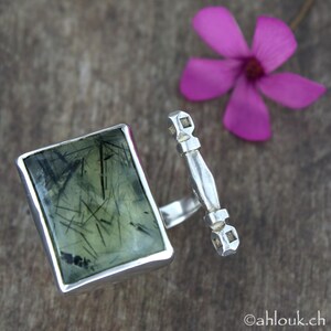 May include: A silver ring featuring a green stone set in a rectangular frame. The stone has a distinctive pattern of black lines running through it. The ring is attached to a silver band with an ornate design.