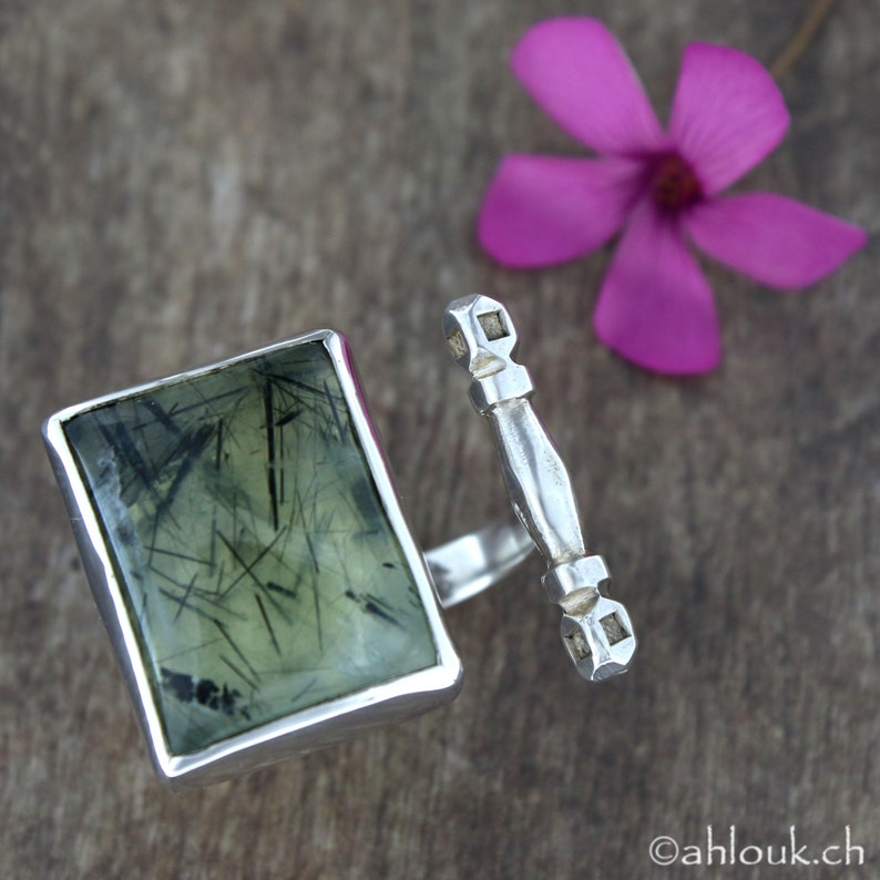 May include: A silver ring featuring a green stone set in a rectangular frame. The stone has a distinctive pattern of black lines running through it. The ring is attached to a silver band with an ornate design.