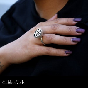 May include: A silver ring with a geometric design. The ring is on a person's finger. The person's nails are painted a dark purple.